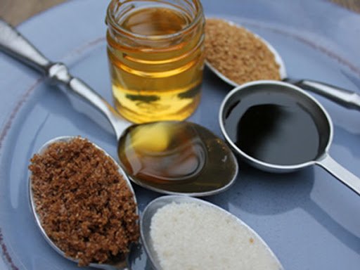 Four spoons with brown sugar, honey, molasses, and white sugar, along with a jar of honey and a jar of raw sugar