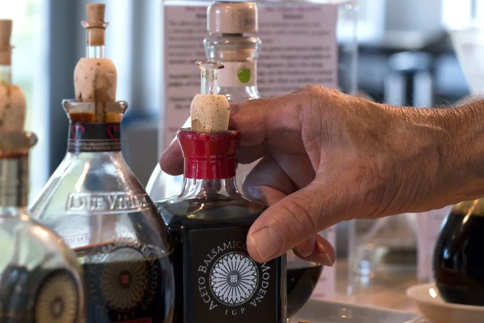 A hand holding a small bottle of balsamic vinegar with a cork stopper and pourer. Other bottles are blurred in the background, indicating a kitchen or store setting.