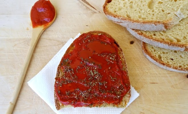 A slice of bread topped with red sauce (Tomato Paste Sauce) and herbs, with a wooden spoon nearby. Additional slices of bread are visible on a wooden cutting board.
