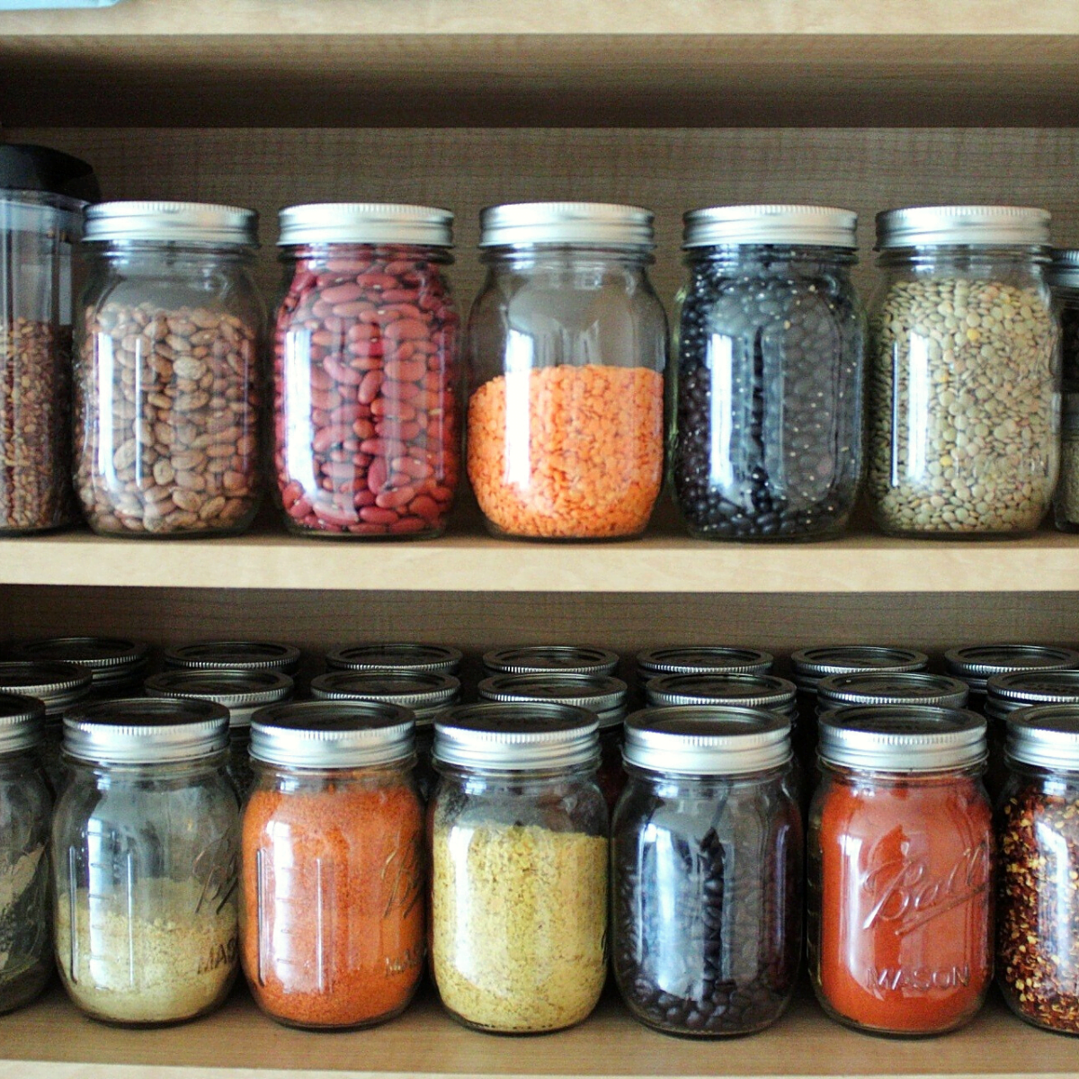 A wooden shelf with neatly arranged glass jars containing a variety of dried goods like beans, lentils, grains, and spices. The jars are well-organized, sealed with lids, and give the impression of a tidy pantry or kitchen storage setup.